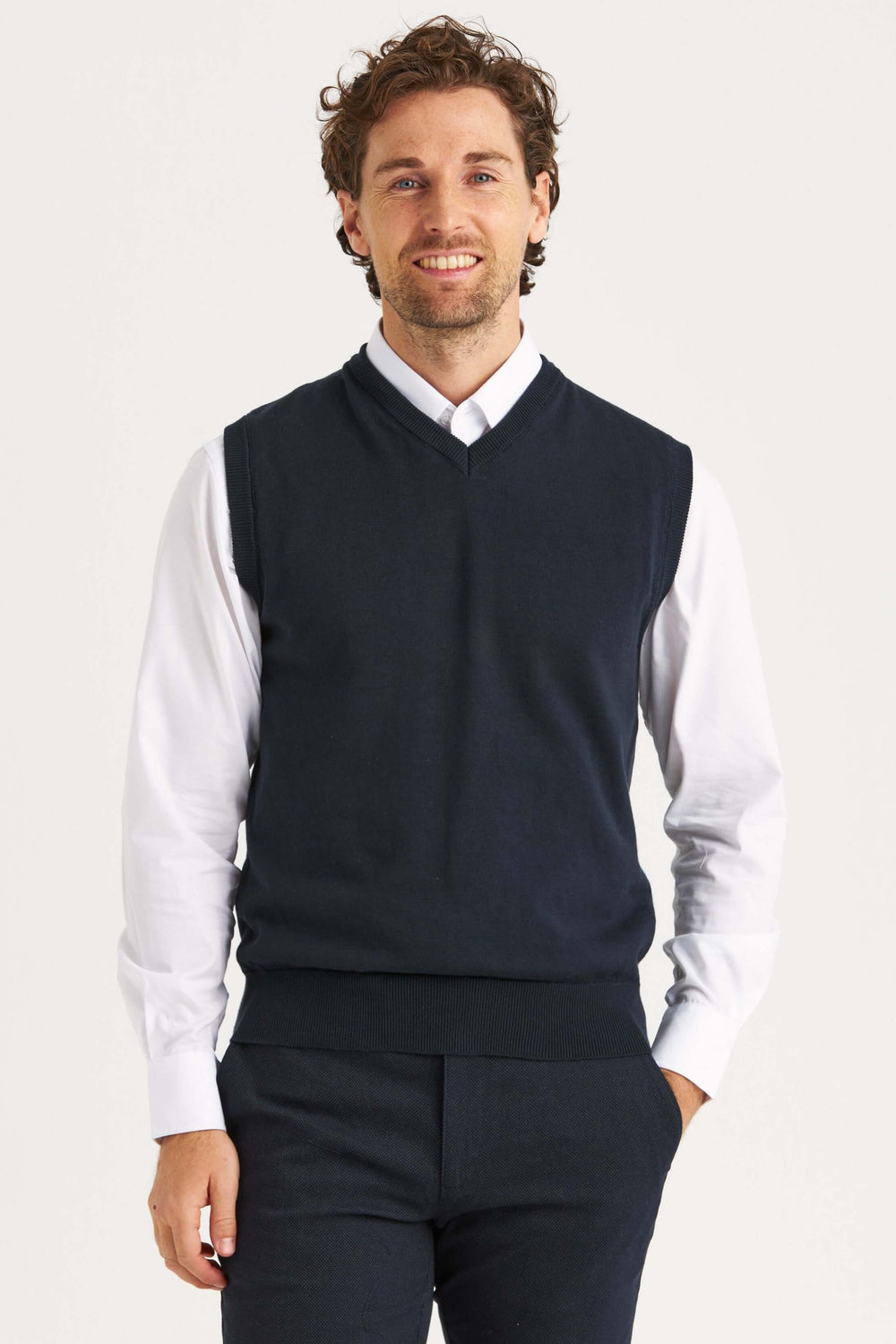 En man bär en marinblå slipover i mjuk bomull över en vit skjorta, perfekt för lager-på-lager look.