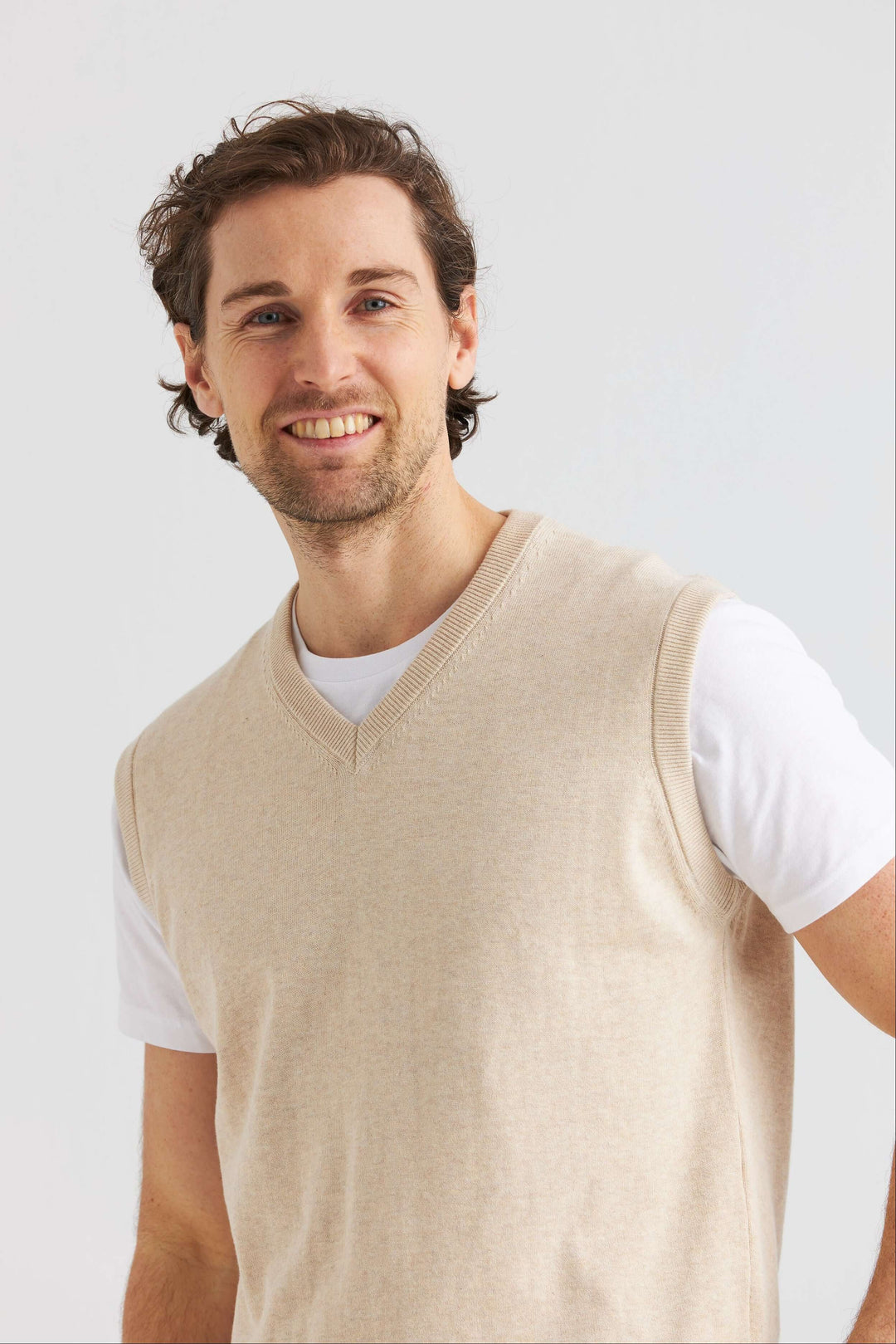 Man i beige slipover i mjuk bomull över vit t-shirt, ler mot kameran på neutral bakgrund.