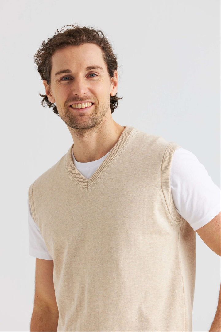 Man i beige slipover i mjuk bomull över vit t-shirt, ler mot kameran på neutral bakgrund.