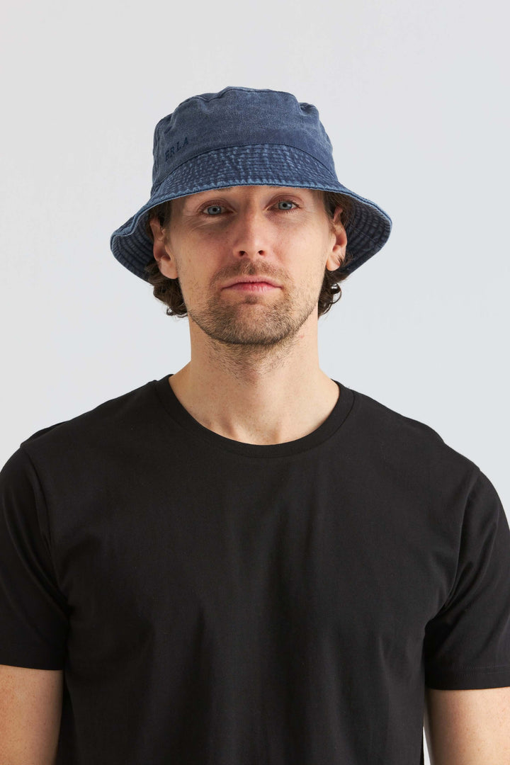 Blå Bucket Hat Washed Cotton bärs av man med svart t-shirt, ser avslappnad och stilren ut.