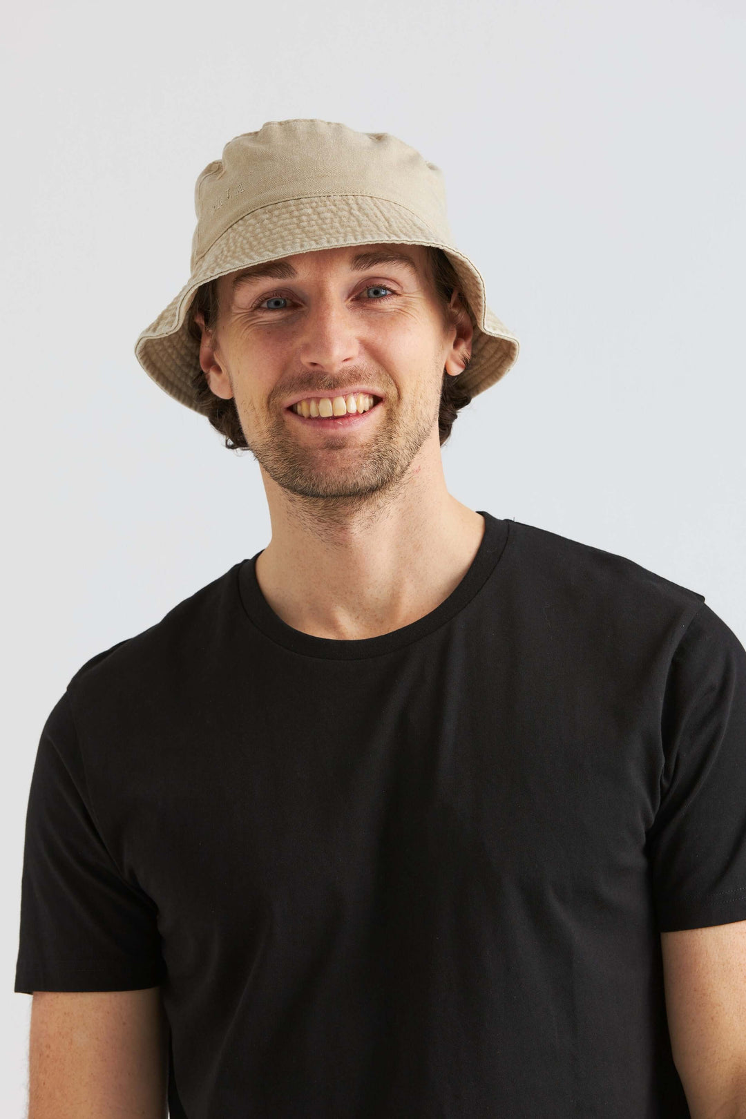 Man wearing a beige bucket hat washed cotton, smiling, with a black t-shirt. Svart Keps Washed Cotton.