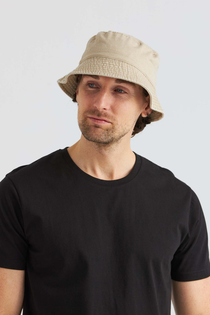 Man wearing a beige bucket hat made of washed cotton, paired with a black T-shirt, stylish summer accessory.
